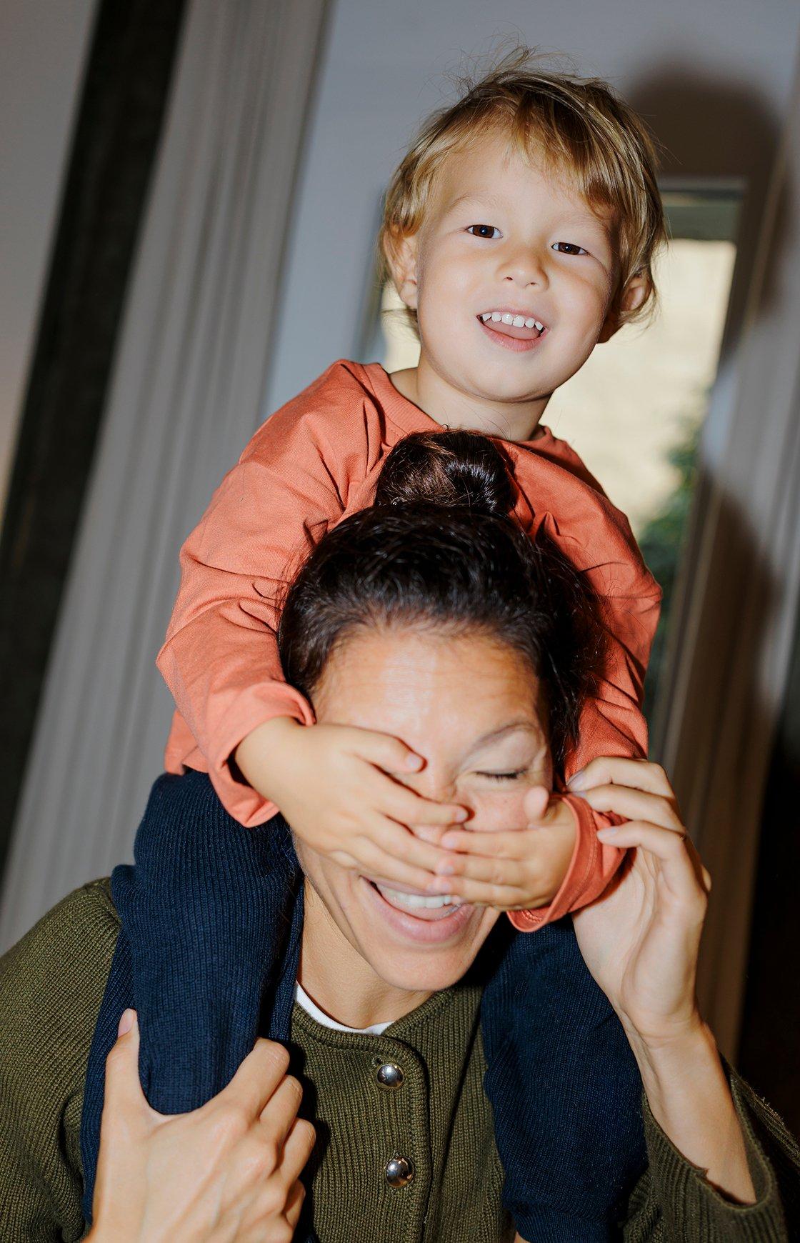 A laughing toddler on his mom's shoulders, covering her eyes with his hands